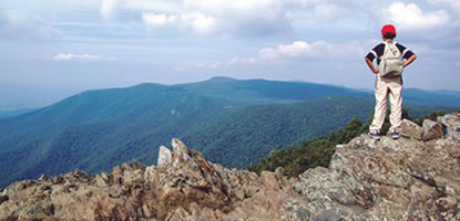 A boy wearing a bright red hat stands on a rocky outcrop and takes in a scenic view.