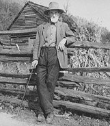 Historic photo of older gentleman in hat with long white beard leaning against a split rail fence.