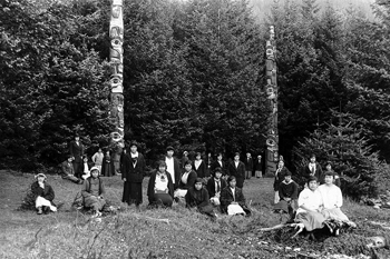 Black and white photograph of a large group of people gathered in a grassy field, with two large totem poles and spruce trees behind them.
