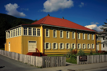 Two-story building painted yellow, with white windows and red roof, surrounded by a wooden picket fence, with blue sky and mountains in the background.