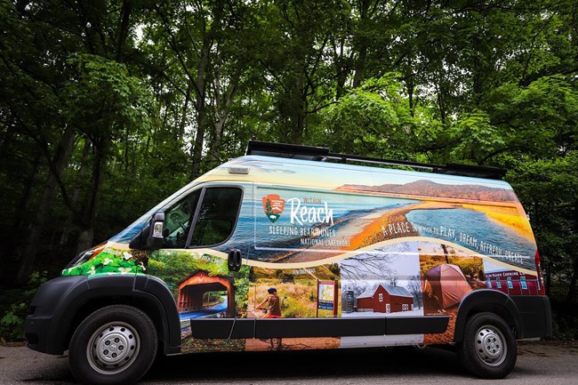 The mobile visitor center van covered in bright images of the park sits along a road with green trees in the background.