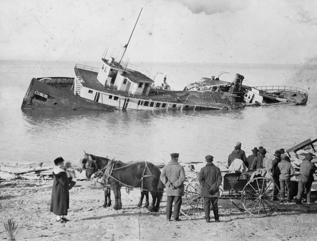 A black and white photo of a partially capsized ship, with watching onlookers and a horse-drawn cart on shore