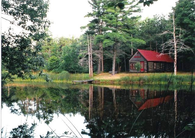 Log cabin with white chinking and red roof, windows, and front door sits on the edge of a pond