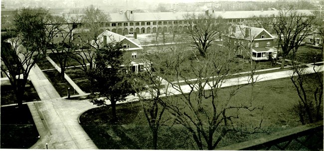 A black and white aerial photo of a long building with multiple windows. Trees and other buildings are in the foreground.