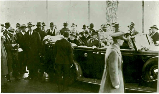 A black and white photo of men crowded around a car with President Roosevelt shaking the hand of John Garand.