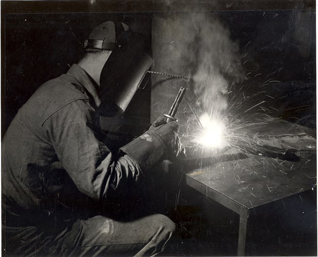 Welding taking place in Building 104. Man at left welds an object on a table in front of him. Man is seated and wears a welder's mask and heavy gloves. Sparks fly from the object.