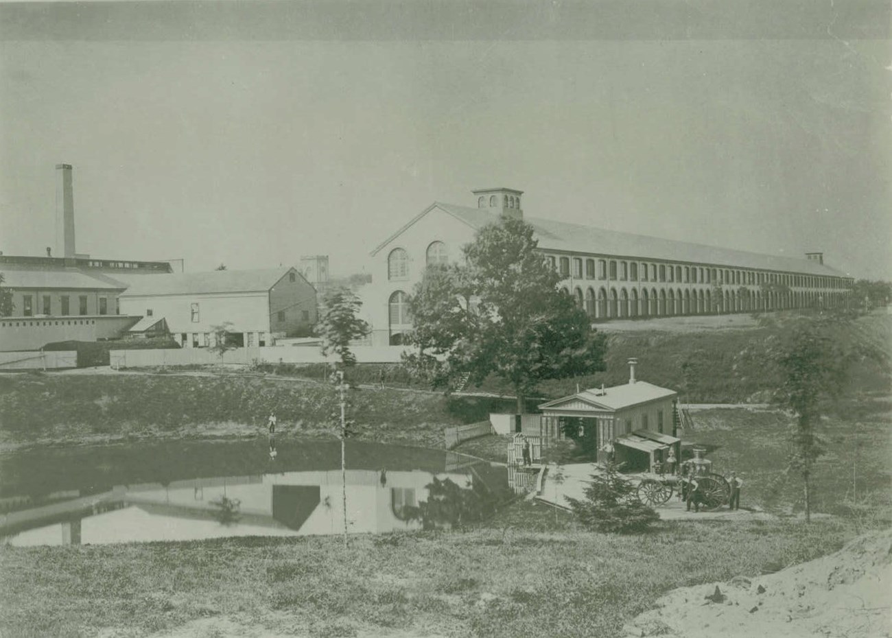 A black and white photo of a long storehouse building with a fire station and pond in the foreground.