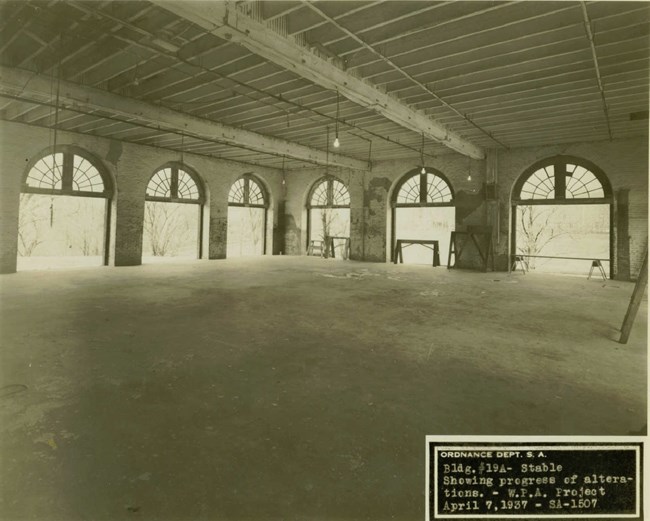 A sepia toned photo of a stables undergoing renovations. There are no stalls, but rather it is a large open room with windows and doors.