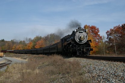 A steam locomotive and passenger excursion train travels along a grassy area in a fall foliage scene.