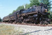 Canadian Pacific 2317 - black with yellow details and a gray front - pulls green passenger cars past trees in the background and gravel in the foreground.