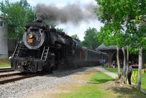 A black steam locomotive pauses near a wooden passenger station during a summer excursion.