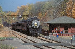 A steam locomotive pulls dark green passenger cars past a brown wooden freight station at Moscow, PA.