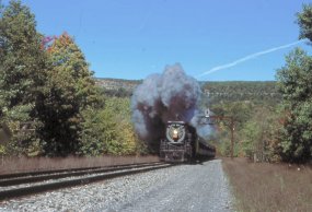 Steam locomotive 3254 puffs gray smoke as it travels under a steel girder signal bridge and passes green forests in Northeastern Pennsylvania.