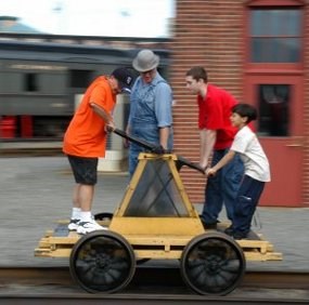 Three children ride on a yellow pump car with a man wearing period overalls and hat.