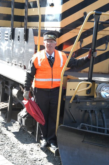 A conductor (the boss of the train) wearing traditional black (but with a modern orange vest for safety) stands on the front steps of the black and yellow Nickel Plate 514 diesel-electric locomotive.