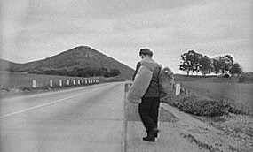 An historic image of a hobo walking along a road with a bed roll slung over his shoulder.