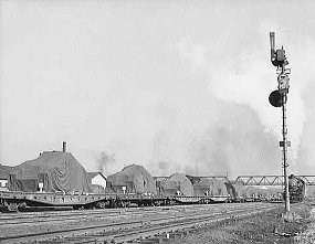 World War II army tanks sit on flat cars covered by tarps in this 1940s photo.