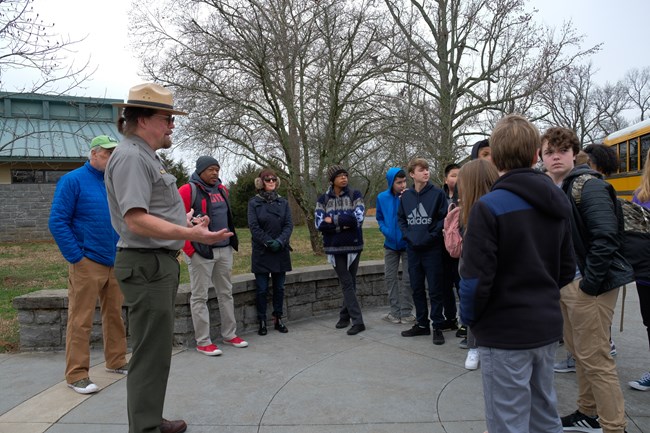 A park ranger speaks to a group of students in front of a school bus.