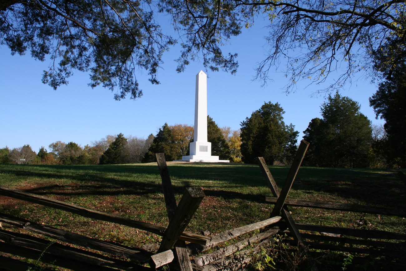 A white obelisk stands on a grass covered hill.