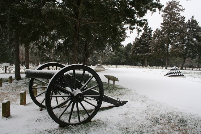 A Civil War cannon sits in a National Cemetery covered in snow, with a pyramid of black cannon balls in the background.