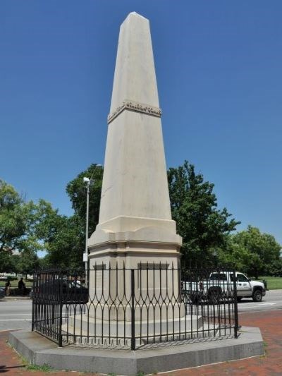 Photograph of a white obelisk monument with a small black fence around the base.