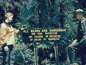 A visitor and a Ranger stand next to a bear warning sign in Glacier National Park, 1958