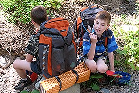 Two little boys wearing backpacks sit and rest