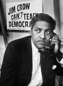 Bayard Rustin on the telephone while sitting in front of a sign reading "Jim Crow Can't Teach Democracy."