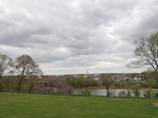 The brick structures and church steeples of a town are seen among spring trees across a river, from atop a grassy area