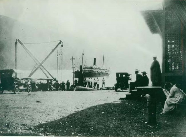 Historical view of people and cars along the waterfront at Kalaupapa Landing, with a large ship in the water.