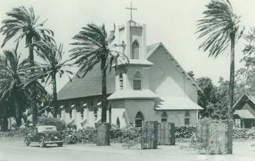 Palms edge a church with a steeple. A smaller structure is visible to the right and a 1930s car is in the road in front.