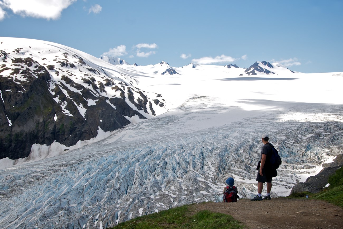 Two people stand next to a dirt trail, looking across Exit Glacier and the Harding Icefield.