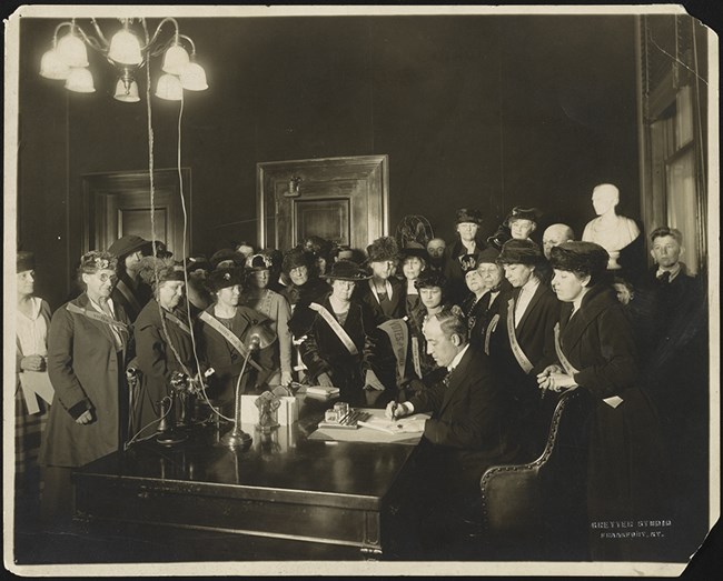 black and white photo of a man at a desk signing a document surrounded by women wearing sashes