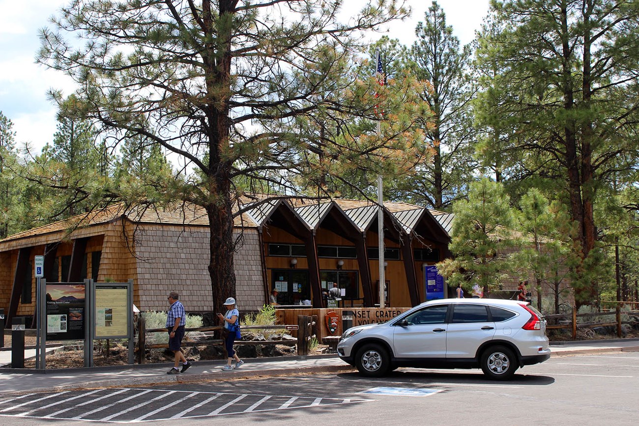 A silver vehicle parked in front of a historic, wooden building with tall columns and a large patio. The building is located in a pine forest.