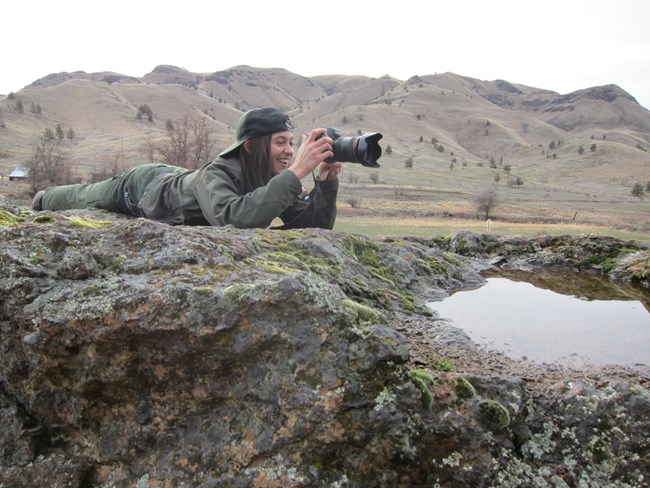 A uniformed Park Ranger smiles as she reviews a photo on a digital camera while laying on a rock.