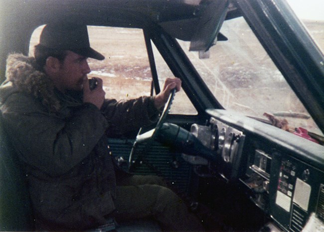 A uniformed man wears a heavy coat and talks on the radio inside a vehicle.