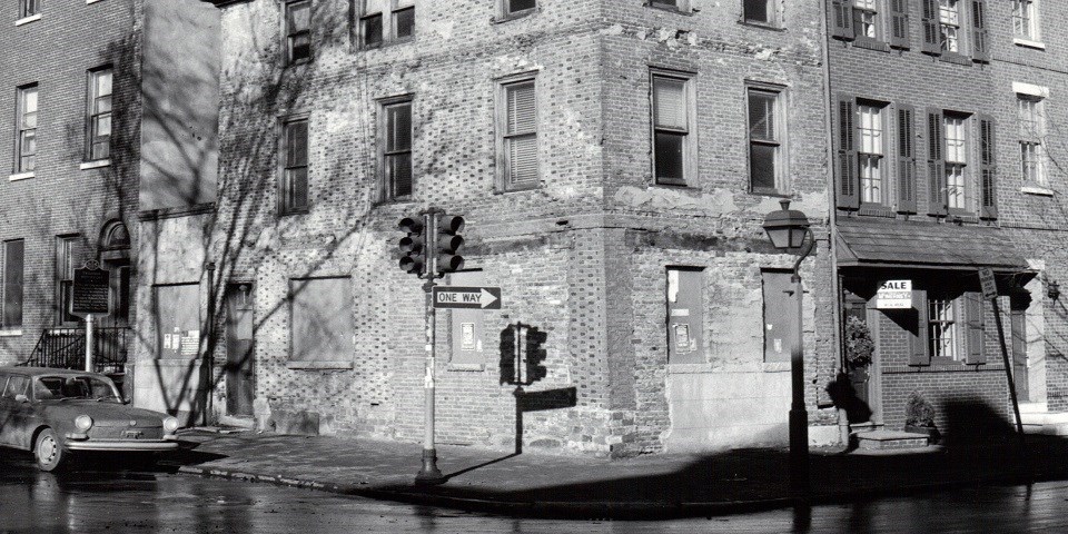 Detail from a black and white photo (circa 1960) showing the exterior of a brick house situated on a corner lot with a car parked next to the building.
