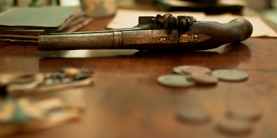 Color photo showing an 18th-century gun lying on a wooden table with coins and a book.