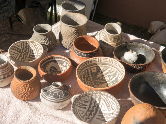 Fourteen pieces of pottery sit on a table. A variety of shapes and styles using red, white, and black pigments.
