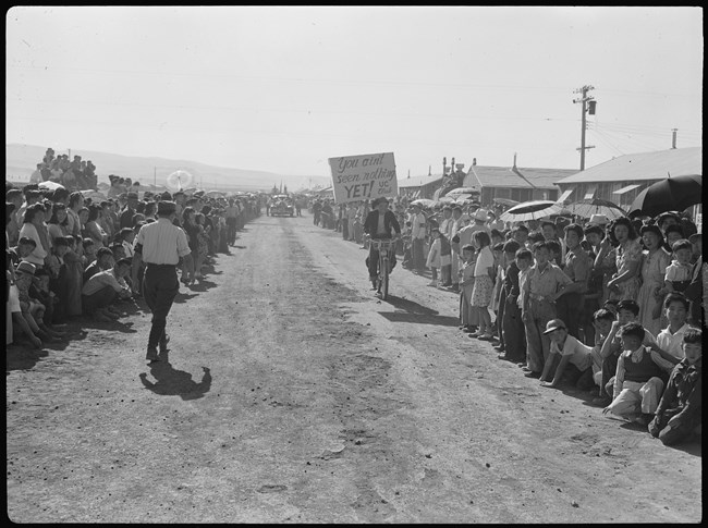Crowds line up on both sides of a dirt road where a man rides a bicycle toward the viewer with a sign that reads "You ain't seen nothing yet! UC Club".
