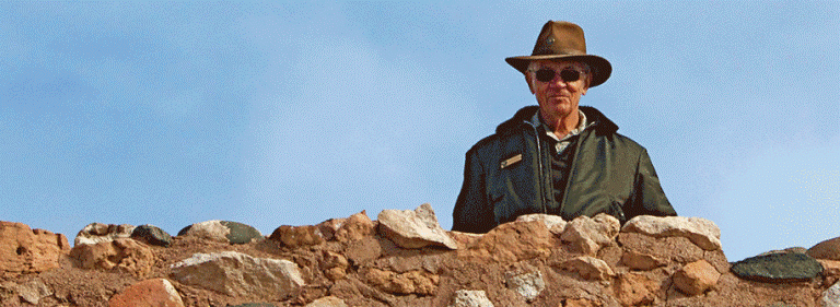 A man in green jacket and hat stands behind a pueblo wall.