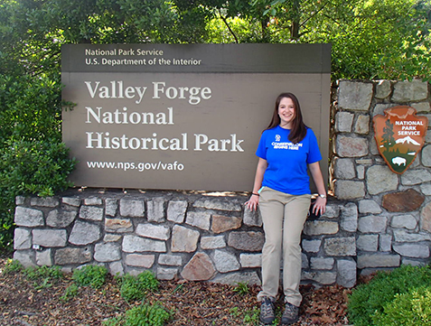 Cristina Ramirez sits on the stone base for the entrance sign to Valley Forge. The National park Service Arrowhead is on her right.