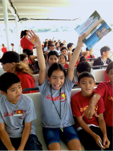 A group of smiling kids on a ferry boat.  The child in front is holding up a Jr. Ranger booklet.