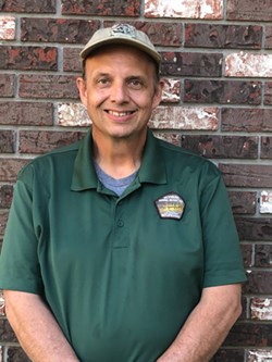 A skinny man smiling with tan hat and green polo shirt.