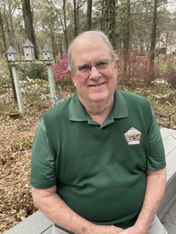 A man sitting in the woods in green polo with glasses smiling.