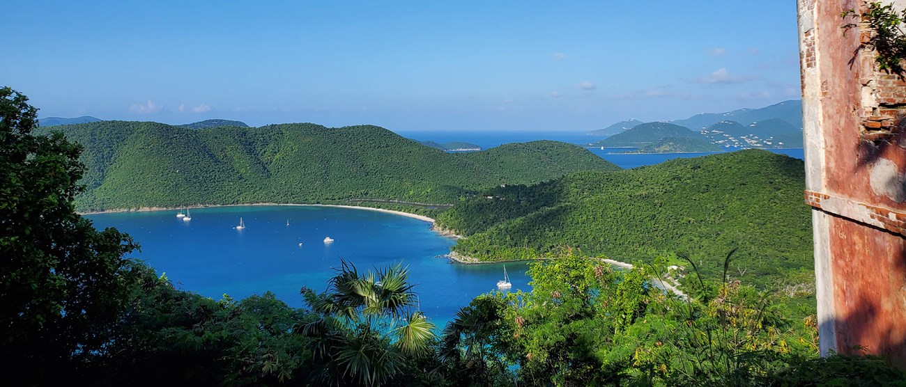 The calm blue water of Francis and Maho Bays are pictured from the America Hill Ruins. St. John's steep hillsides are covered in thick green vegetation, and the BVI can be seen in the distance.
