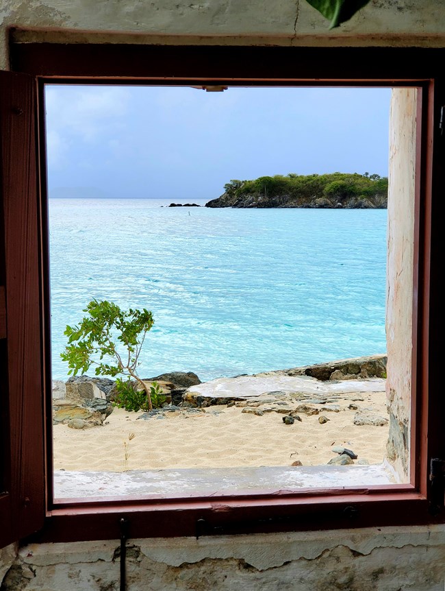 The rocky shoreline and green vegetation of Cinnamon Cay are pictured through one of the windows of the old Danish warehouse on Cinnamon Bay Beach with it's white sand and turquoise waters.
