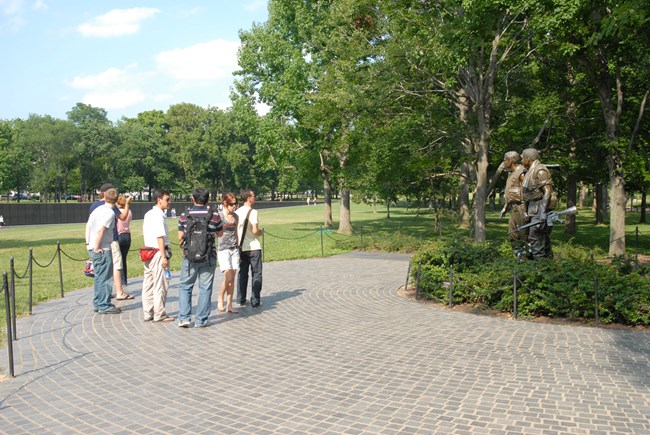 Visitors stand in front of the Three Servicemen Statue. The Vietnam Veterans Memorial wall stands in the beackground.