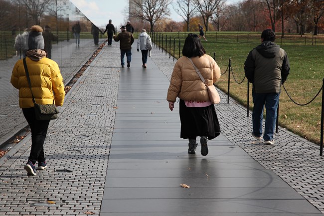 Visitors walk along the memorial's pathway, which is an ascending brick and granite surface. The Washington Monument is peeking through trees in the background.