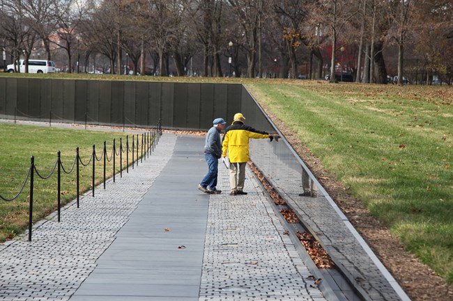 A park volunteer points to a name on the memorial wall as a visitor peers at the name.
